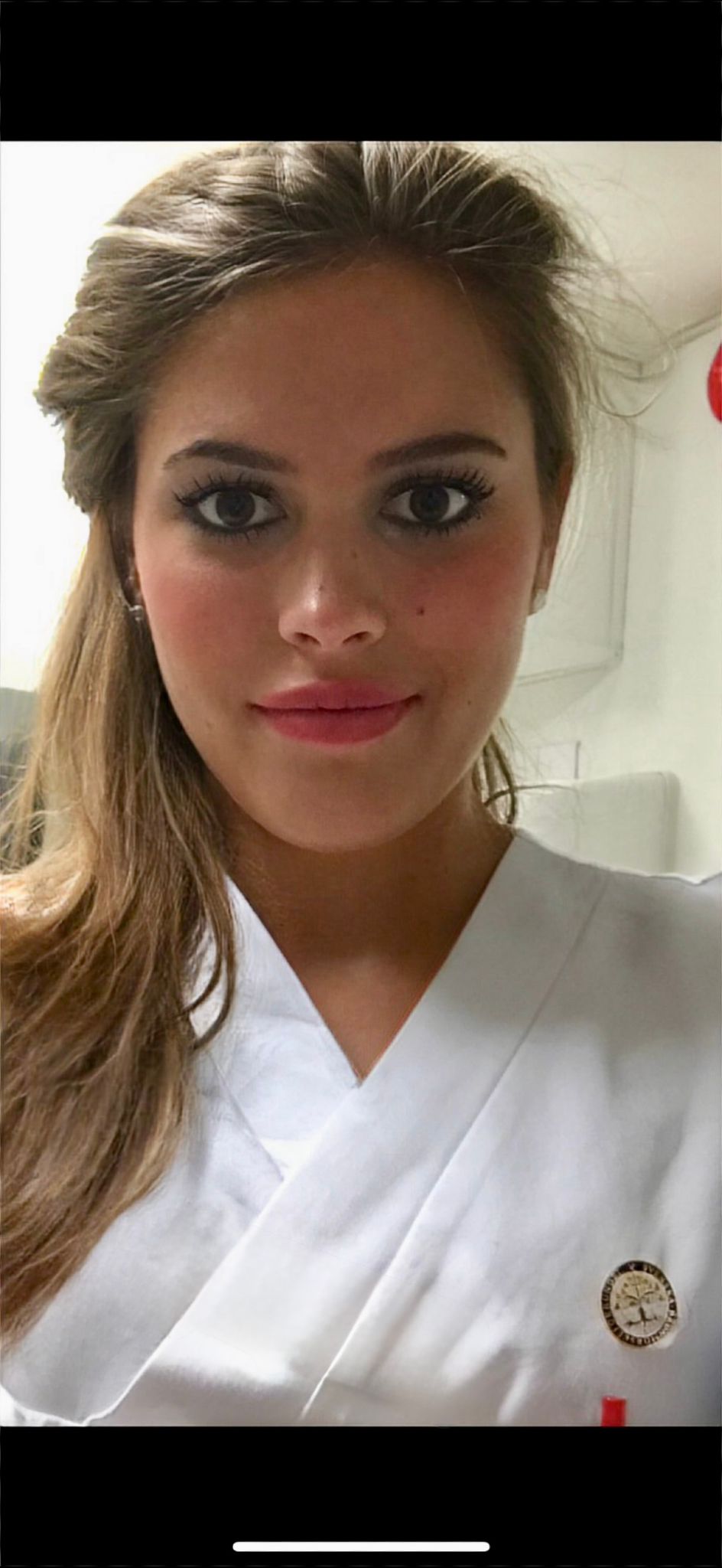 En ung kvinna med långt brunt hår, klädd i vit uniform och med en rund nål, tittar in i kameran med ett litet leende. Bakgrunden ser ut att vara en ljus inomhusmiljö.