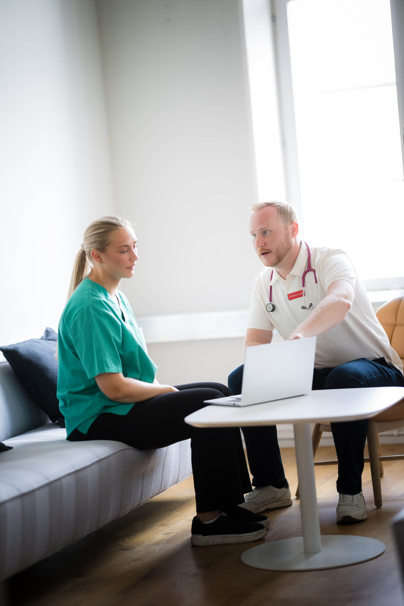 En läkare med stetoskop diskuterar något på en bärbar dator med en sjuksköterska som sitter i en soffa på en ljus, modern läkarmottagning.