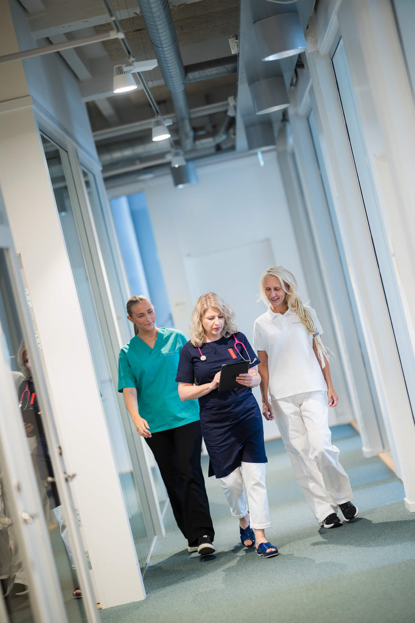 Tre vårdpersonal går längs en korridor på en vårdinrättning. En av dem har gröna operationskläder, en annan marinblå och den tredje vita. De ser ut att vara inbegripna i en konversation.