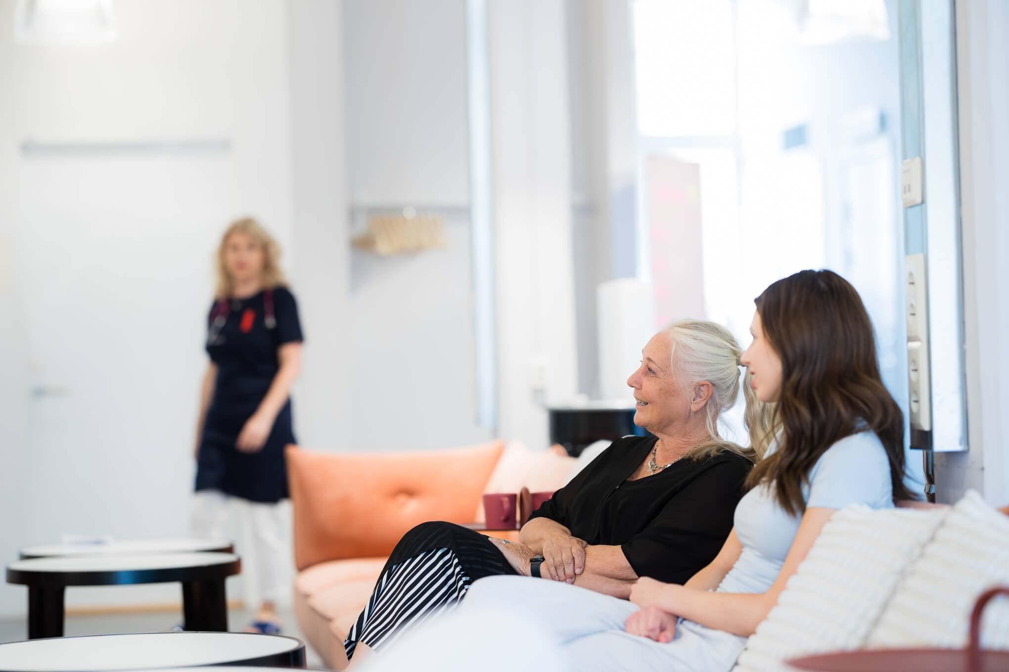 Två kvinnor sitter i en soffa i ett väntrum och pratar och ler. En person i läkarkläder går mot dem i bakgrunden. Miljön verkar lugn och väl upplyst.