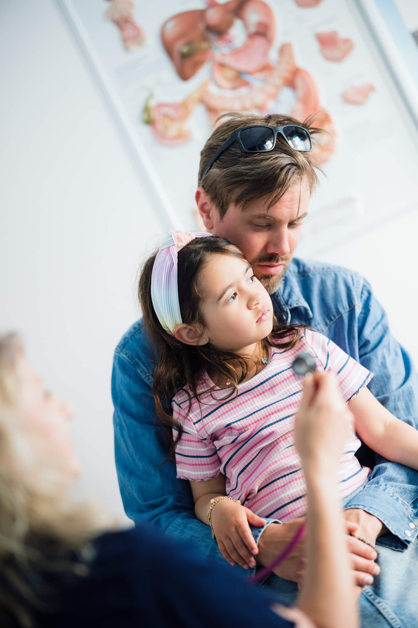 En man håller en ung flicka i sitt knä medan en kvinna, troligen en läkare, håller ett stetoskop mot flickan. Medicinska diagram syns på väggen i bakgrunden.
